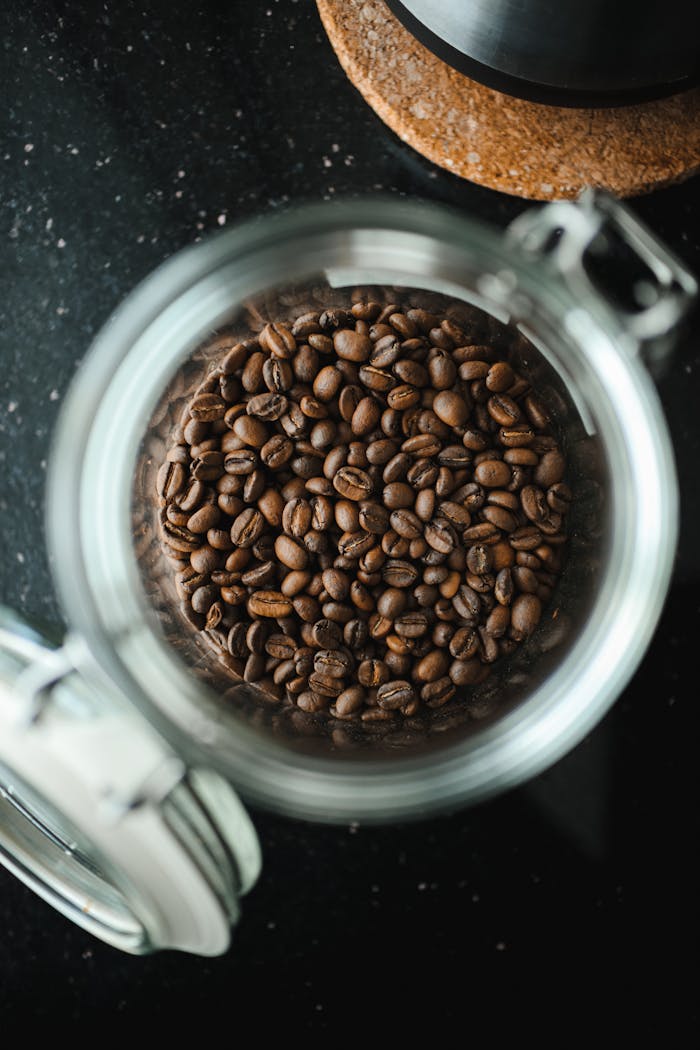 Top view of roasted coffee beans in an open glass jar on a dark surface.